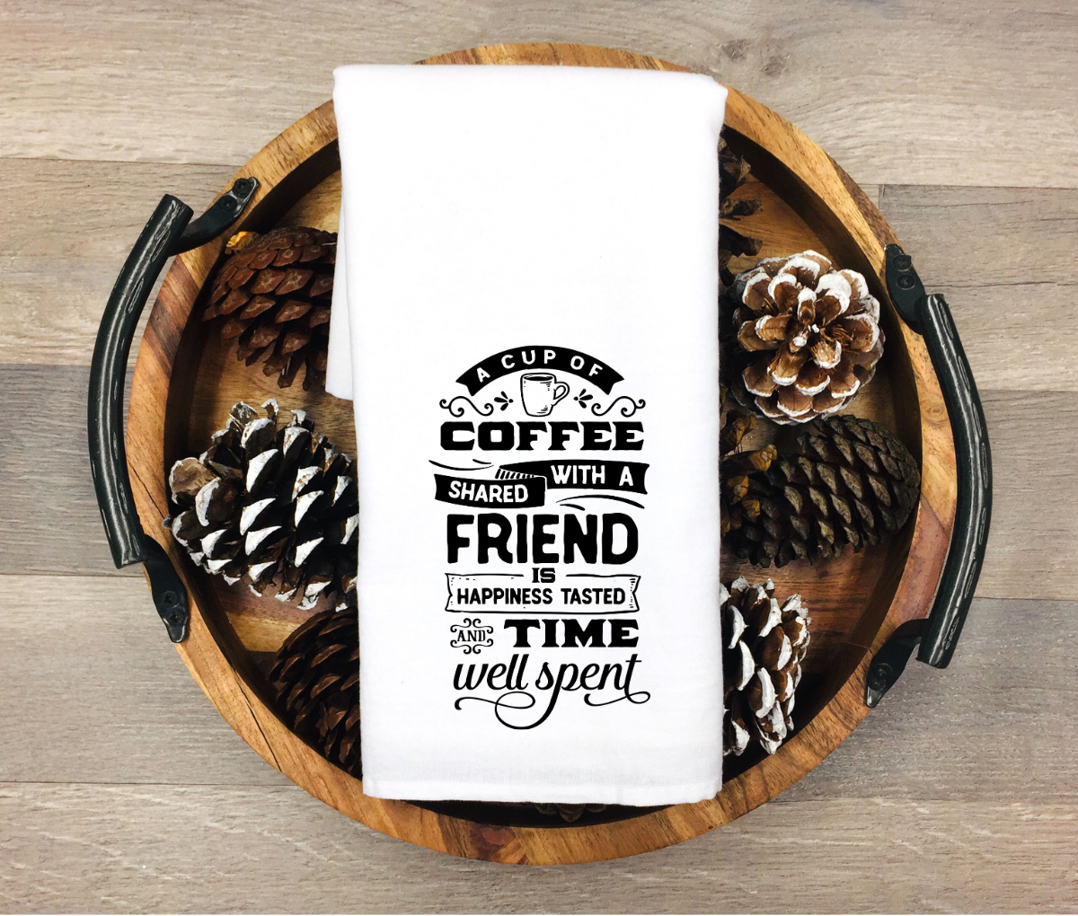 White kitchen towel featuring the quote “A cup of coffee shared with a friend is happiness tasted and time well spent” in black decorative typography, displayed on a wooden tray with pinecones.