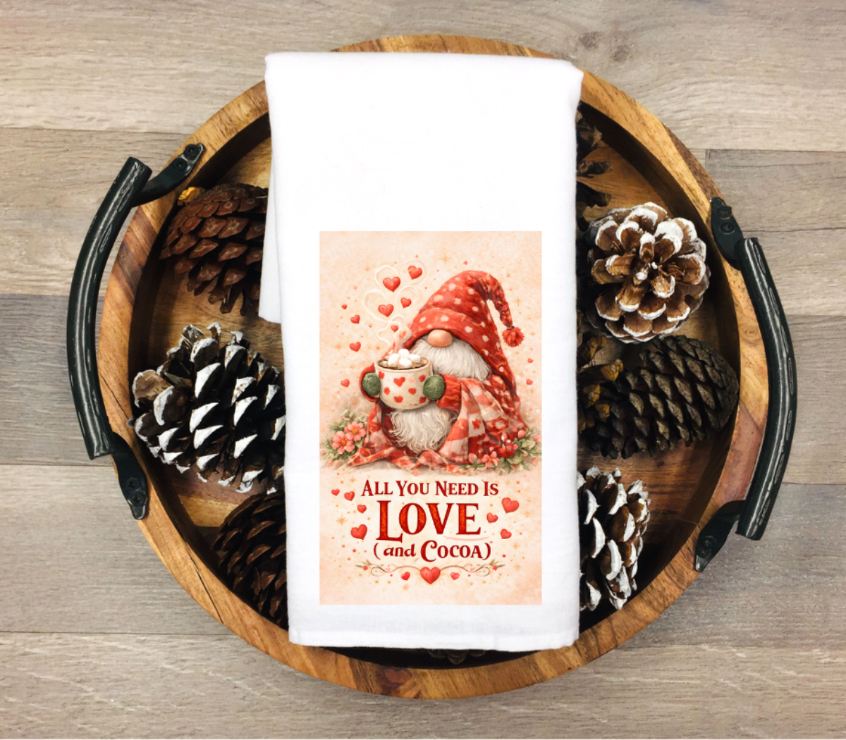 White kitchen towel with a cozy gnome holding a mug of hot cocoa and the quote “All You Need Is Love (and Cocoa)” in red lettering.