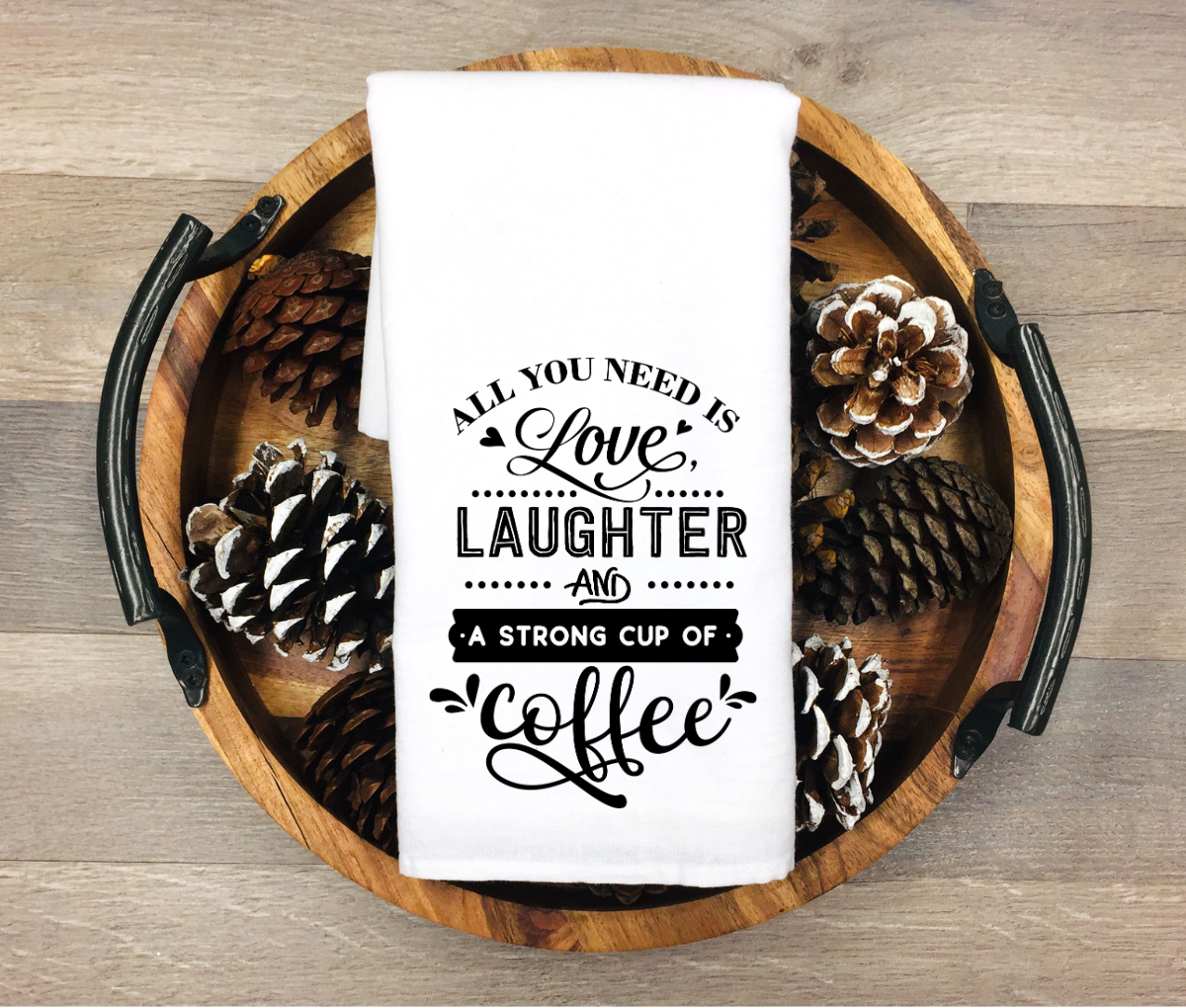 White towel with 'All you need is love, laughter, and a strong cup of coffee' text on a wooden tray with pinecones.