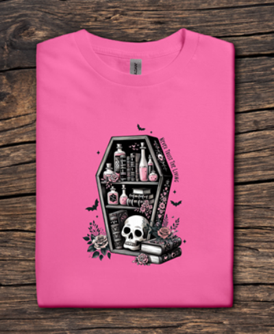 Pink t-shirt with a black coffin design featuring a skull and various items on a wooden surface