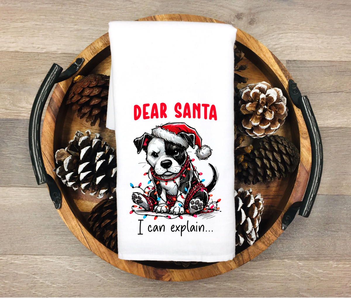 White towel with Christmas-themed design featuring a dog and text, placed on a wooden tray with pinecones.