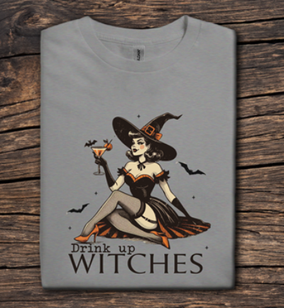 Gray t-shirt with a witch graphic and 'Drink up Witches' text on a wooden surface