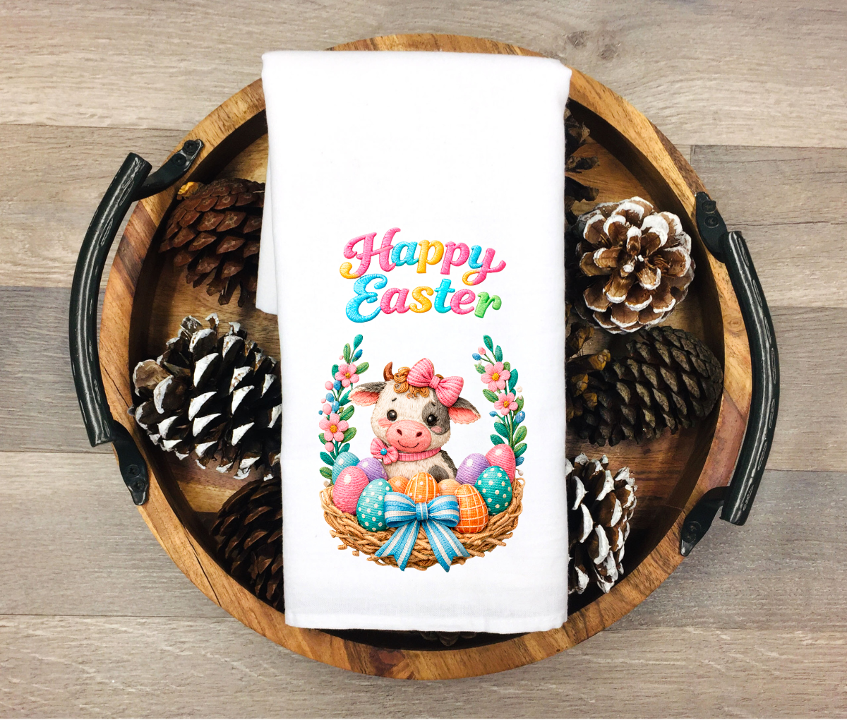 Easter-themed towel with a cow and eggs design on a wooden tray with pinecones.