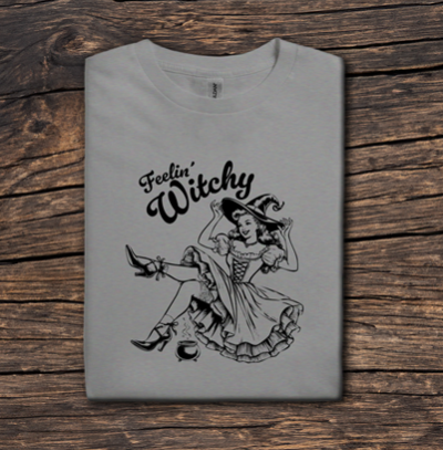 Gray t-shirt with 'Feelin' Witchy' design on a wooden surface