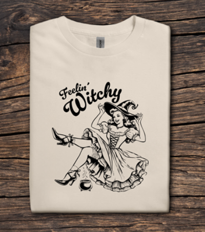 Beige t-shirt with 'Feelin' Witchy' graphic design on a wooden surface