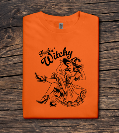 Orange t-shirt with a witch graphic and 'Feelin' Witchy' text on a wooden surface