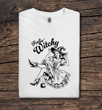 White cotton t-shirt featuring a vintage-style illustration of a playful witch sitting beside a bubbling cauldron with the phrase “Feelin’ Witchy” in bold script above her.