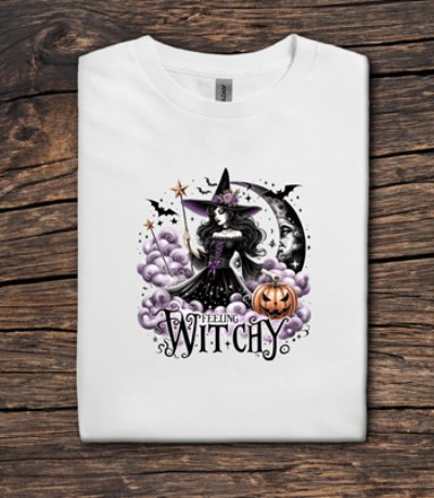 White t-shirt with Halloween-themed design featuring a witch, moon, and pumpkin on a wooden surface.