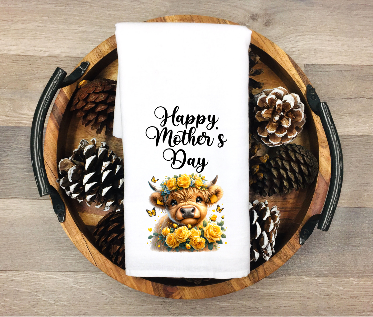 White towel with 'Happy Mother's Day' text and cow illustration on a wooden tray with pinecones.