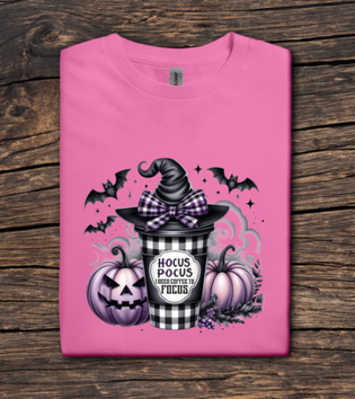 Pink t-shirt with Halloween-themed design featuring a witch hat, pumpkins, and 'Hocus Pocus' text on a wooden surface.
