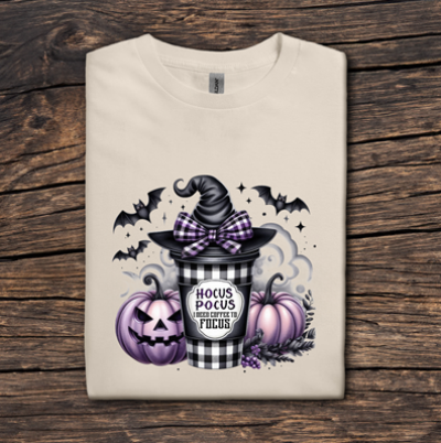 Beige t-shirt with Halloween-themed design featuring a witch hat, pumpkins, and text on a wooden surface.