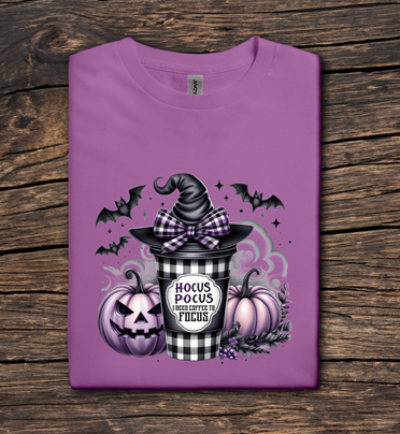 Purple t-shirt with Halloween-themed design featuring a witch hat, pumpkins, and text on a wooden surface.