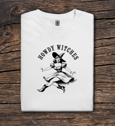 White t-shirt with a graphic of a witch and 'Howdy Witches' text on a wooden surface