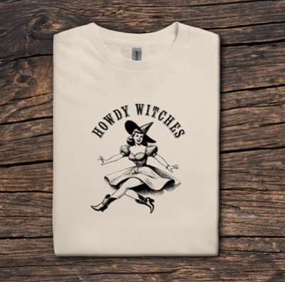 Beige t-shirt with 'Howdy Witches' graphic on a wooden surface