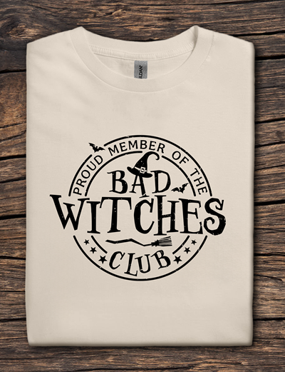 Beige t-shirt with 'Proud Member of the Bad Witches Club' text on a wooden surface