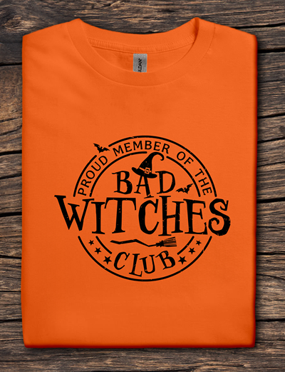 Orange t-shirt with 'Proud Member of the Bad Witches Club' text on a wooden surface