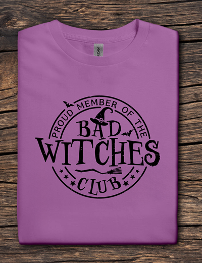 Purple t-shirt with 'Proud Member of the Bad Witches Club' text on a wooden surface