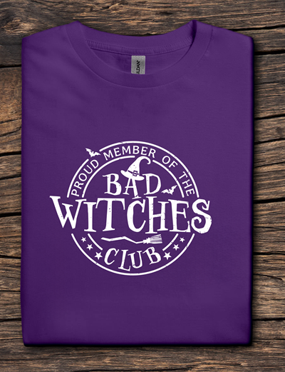 Purple t-shirt with 'Proud Member of the Bad Witches Club' text on a wooden surface
