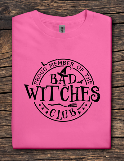 Pink t-shirt with 'Proud Member of the Bad Witches Club' text on a wooden surface